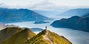 Person standing on a mountain peak overlooking a lake with mountains in the background